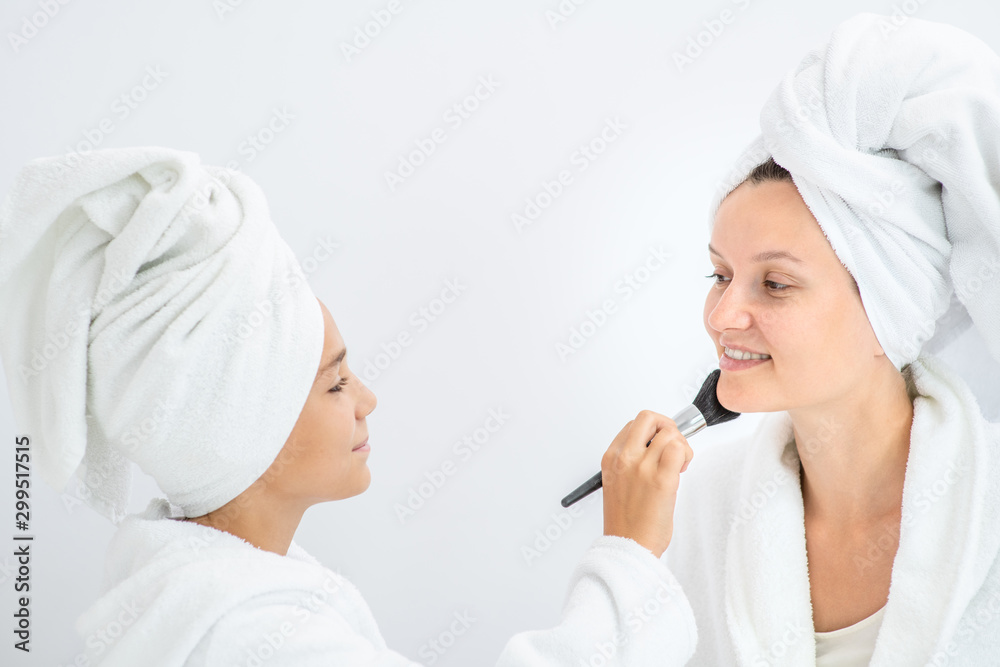 Young girl applying powder on nose her mother using a brush. Mom and child girl are in bathrobes and with towels on their heads Young girl applying powder on nose her mother using a brush. Mom and child girl are in bathrobes and with towels on their heads