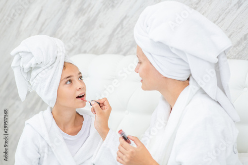 Happy family at home. mom paints her daughter's lips on the bed at home. Mom and girl are in bathrobes and with towels on their heads