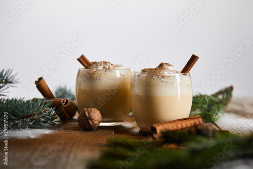 selective focus of eggnog cocktail with whipped cream near cinnamon sticks and spruce branches on wooden table isolated on grey photo