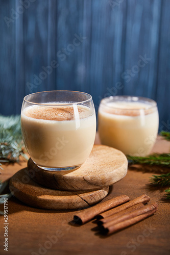 delicious eggnog cocktail on round wooden boards near spruce branches and cinnamon sticks on blue textured background