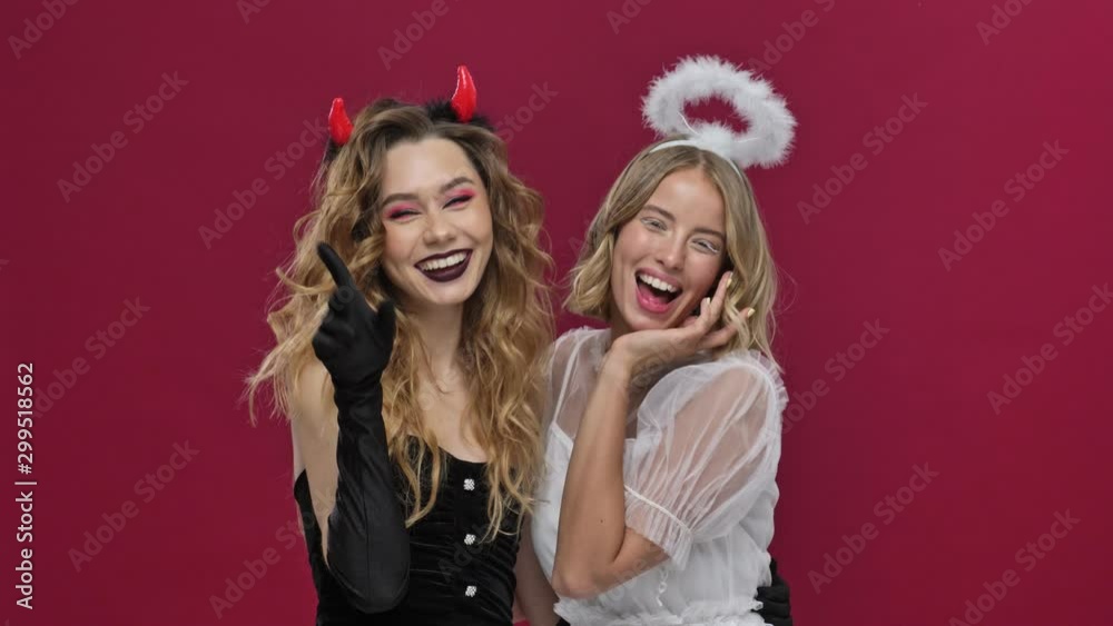Angel and demon girls send kisses at the camera smile and laugh in carnival costumes isolated over red wall background Angel and demon girls send kisses at the camera smile and laugh in carnival costumes isolated over red wall background