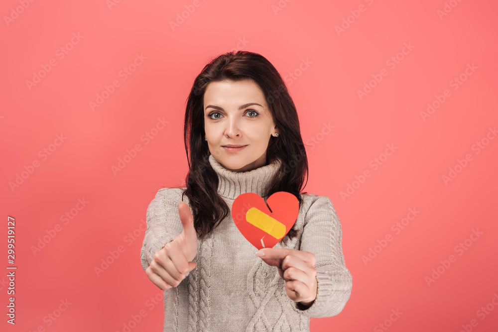 woman in warm sweater holding paper cut with broken heart and band aid while showing thumb up on pink woman in warm sweater holding paper cut with broken heart and band aid while showing thumb up on pink