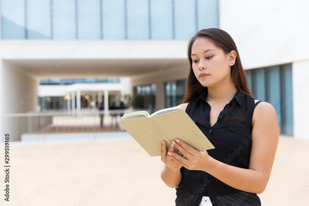 Focused female student reading notes or book. Young Latin woman standing near city building, holding open notebook. Education concept Focused female student reading notes or book. Young Latin woman standing near city building, holding open notebook. Education concept