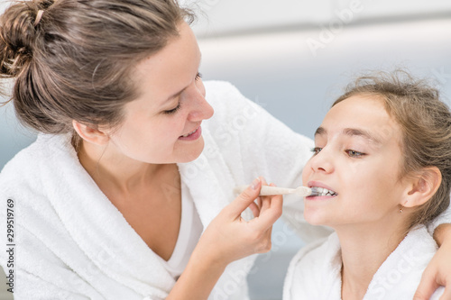 Smiling mom teaching her young daughter how to brush teeth with toothbrush