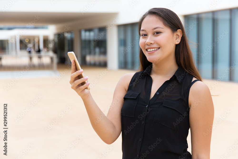 Cheerful smiling student girl using online app. Young mix raced woman standing outside, holding mobile phone, looking at camera. Wireless internet connection concept Cheerful smiling student girl using online app. Young mix raced woman standing outside, holding mobile phone, looking at camera. Wireless internet connection concept