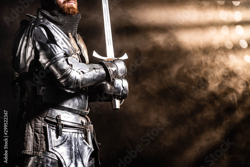 cropped view of knight in armor holding sword on black background photo
