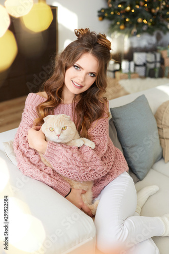 Christmas portrait of a happy young woman in cozy sweater hugging fluffy cat in the interior decorated for New year party. Beautiful model girl in winter outfit hugging a cute kitty and smiling.