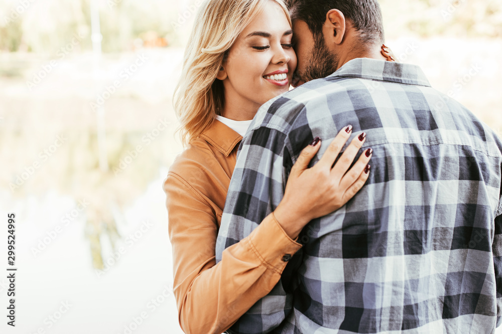 Fototapeta premium happy young woman embracing boyfriend while standing near lake in park