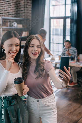 cheerful businesswoman sticking out tongue while taking selfie on smartphone with cheerful colleague winking and holding coffee to go