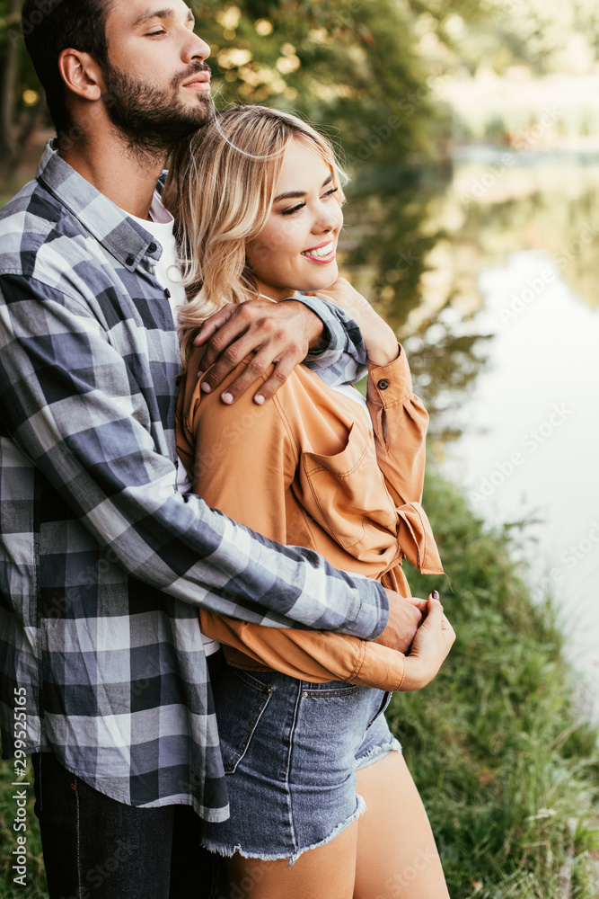 handsome man embracing smiling girlfriend while standing near lake in park handsome man embracing smiling girlfriend while standing near lake in park