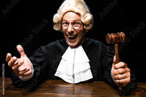Plakat w ramie smiling judge in judicial robe and wig sitting at table and holding gavel isolated on black