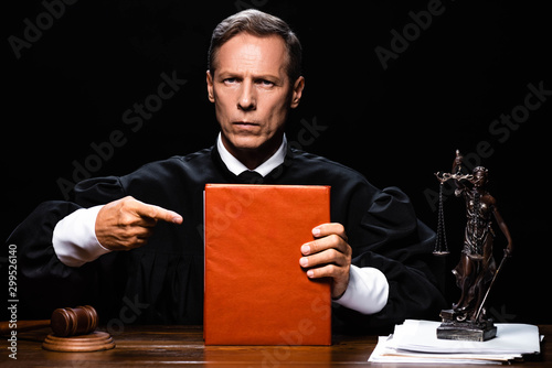 Leinwand Poster judge in judicial robe sitting at table and pointing with finger at orange book