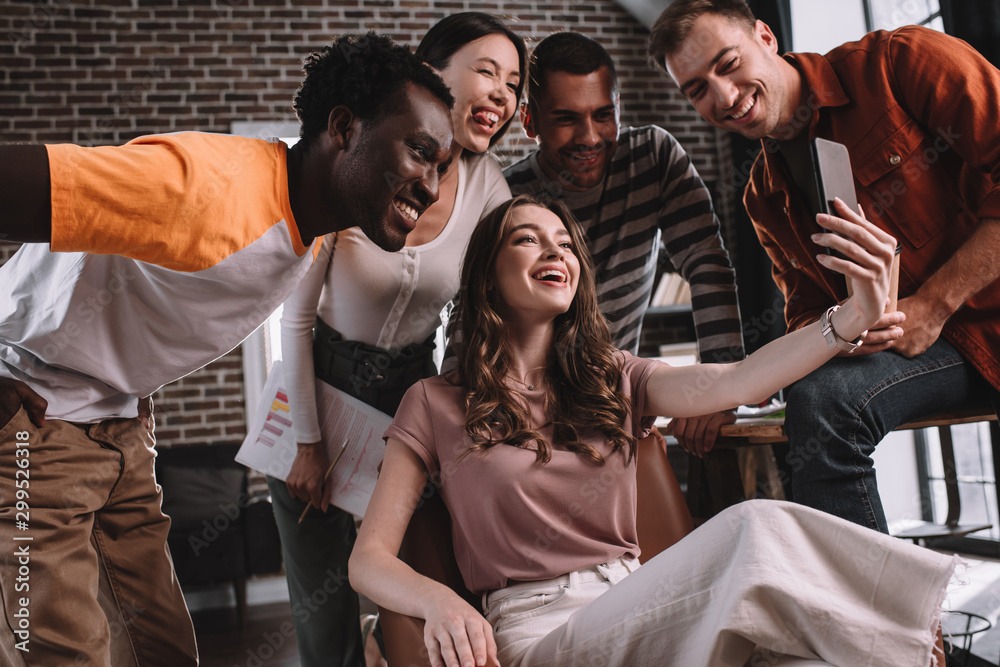 Fototapeta premium happy businesswoman taking selfie on smartphone while sitting in armchair surrounded by cheerful multicultural colleagues