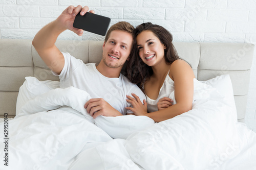young couple handsome husband and his beautiful wife just woke up in bed shooting selfie on smartphone early in the morning social media addiction photo