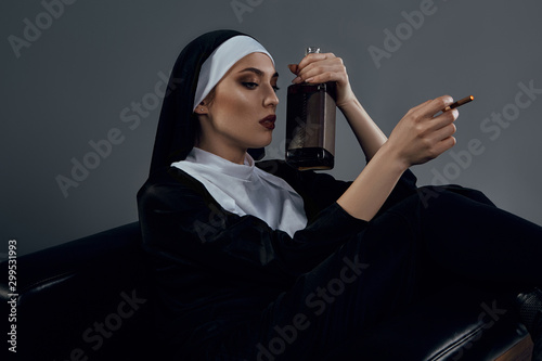 Cropped half-turn shot of a nun, sitting on a black chair. She's wearing dark nun's clothing. The nun is smoking and holding bottle of whiskey in left hand. 