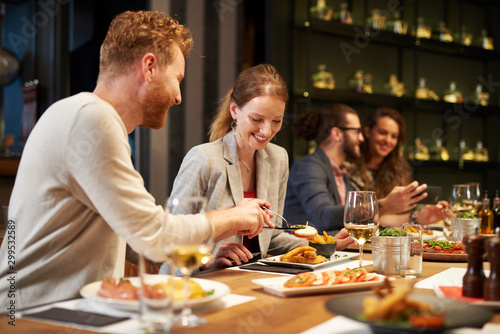 Handsome caucasian ginger taking food out of his girlfriend's plate while sitting in restaurant for dinner. In background are their friends. © chika_milan