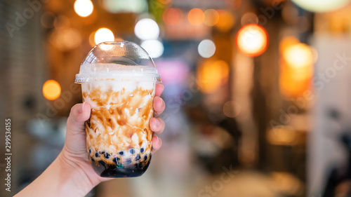 Young woman is holding, drinking brown sugar flavored tapioca pearl bubble milk tea with glass straw in night market of Taiwan, close up, bokeh