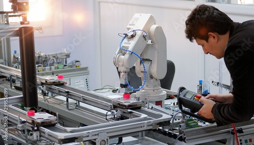 Man is holding teach panel to control a robot arm which is integrated on smart factory production line. industry 4.0 automation line which is equipped with sensors and robotic arm. Selective Focus.