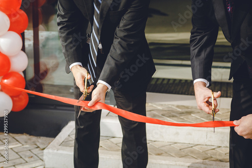 Businessman cutting red ribbon with pair of scissors. Two man in a classic black official suit cuts a red tape opening place