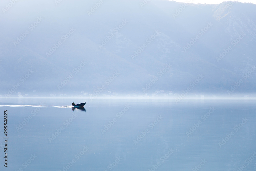 Fototapeta premium Blue nature background. Boat with fisherman on sea. Fishing in foggy morning lake. Amazing seaside landscape with mountains, silence, calmness. Reflection in still water of the Kotor Bay, Montenegro.