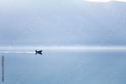 Blue nature background. Boat with fisherman on sea. Fishing in foggy morning lake. Amazing seaside landscape with mountains, silence, calmness. Reflection in still water of the Kotor Bay, Montenegro.
