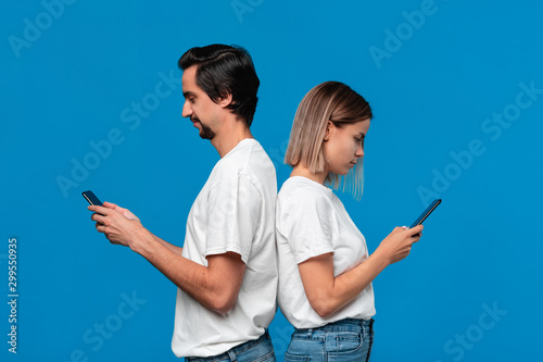 Blond woman and brunet man in white tess and blue jeans looking at screens of their mobile phones standing with their back to each other. photo