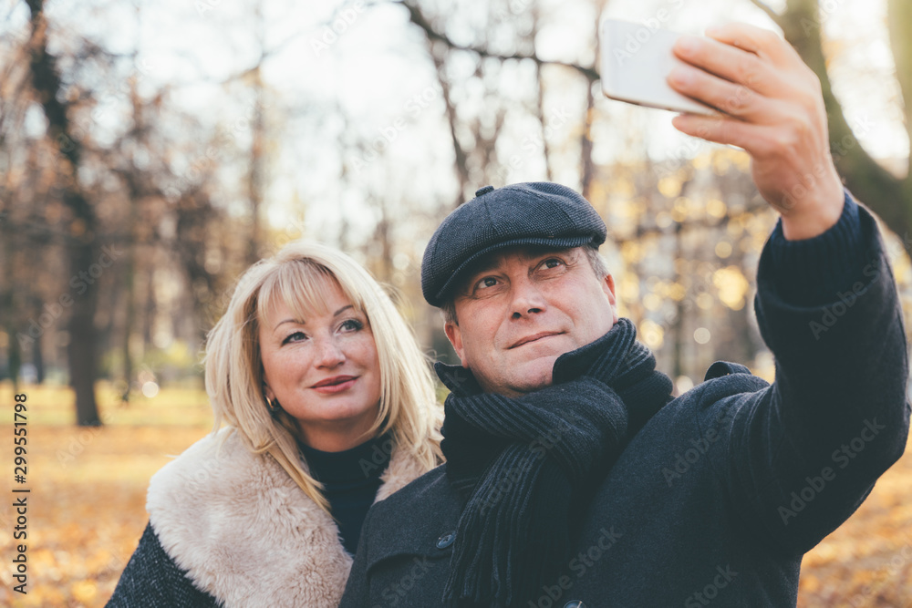 Fototapeta premium Happy blonde mature woman and beautiful middle-aged brunette take selfie on mobile phone. Loving couple of 45-50 years old walks in autumn park in warm clothes, in coat and enjoys life
