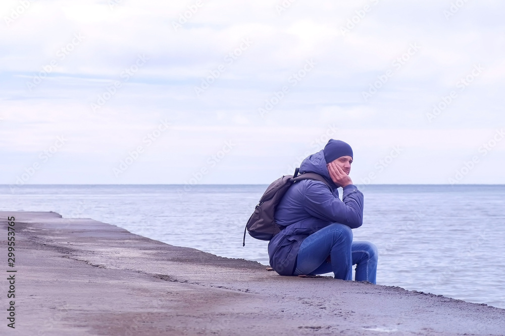 Tourist at sea off-season. He is wearing on hat, jacket, jeans with backpack. Man traveller sits on old waterfront at sea in winter day and looks at seascape. Tourist at sea off-season. He is wearing on hat, jacket, jeans with backpack. Man traveller sits on old waterfront at sea in winter day and looks at seascape.