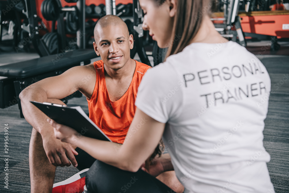 personal trainer showing clipboard to smiling african american sportsman sitting on floor