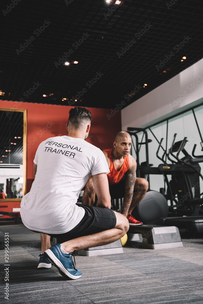Obraz premium back view of personal trainer supervising african american athlete lifting weight in gym
