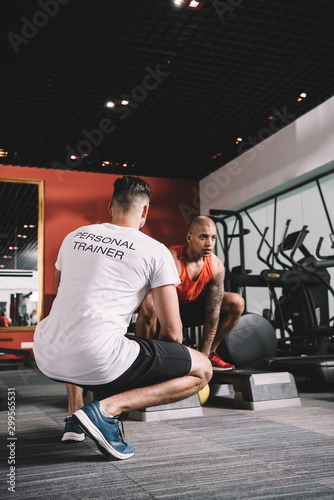 back view of personal trainer supervising african american athlete lifting weight in gym