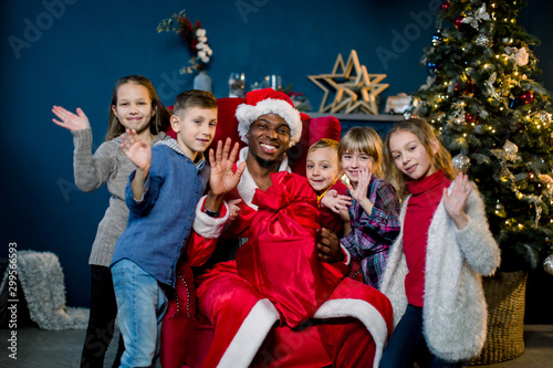 Beautiful five children, three girls and two boys, laughing, hugging African Santa Claus, waving their hands. Christmas holidays, New Year