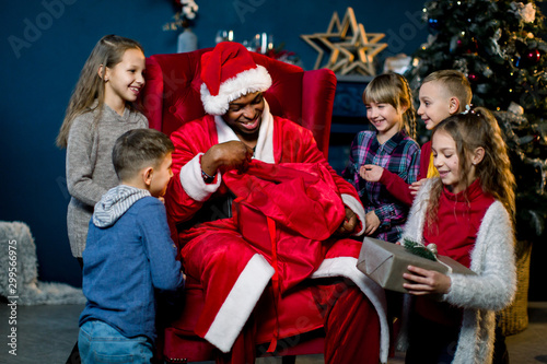 African Santa Claus sitting in armchair and giving the presents to beautiful little children in a christmas-decorated room. New Year, winter holidys, Christmas concept photo