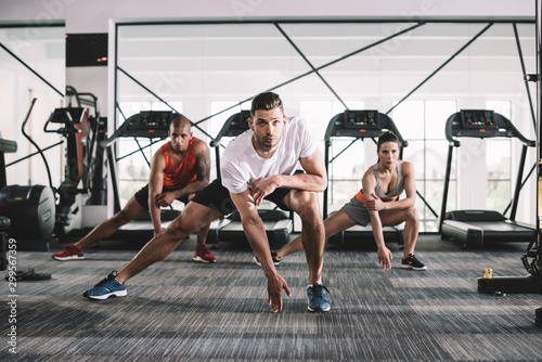 handsome trainer looking at camera while doing lunges exercise together with multicultural athletes