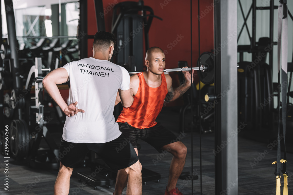 Fototapeta premium back view of personal trainer controlling young african american sportsman lifting barbell