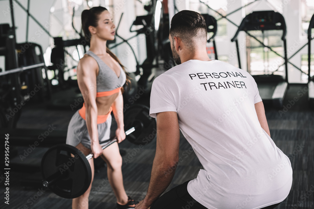 back view of personal trainer looking at attractive sportswoman lifting barbell back view of personal trainer looking at attractive sportswoman lifting barbell