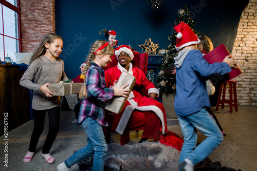 African Santa Claus is seated in red armchair. Happy children run with Santa Claus gifts in the christmas room