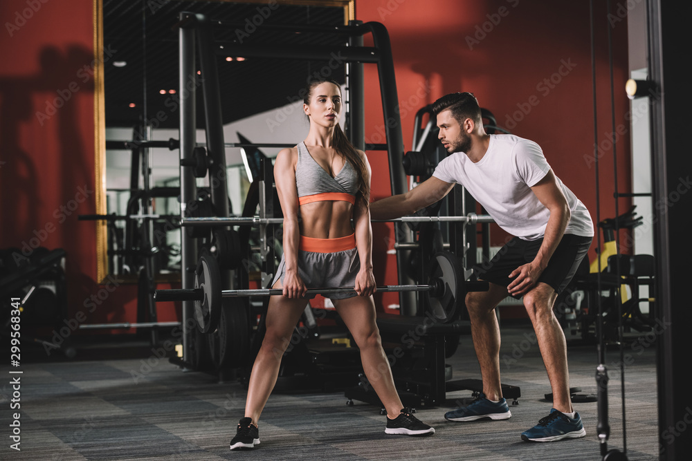 handsome trainer supporting attractive sportswoman lifting barbell - obrazy, fototapety, plakaty 