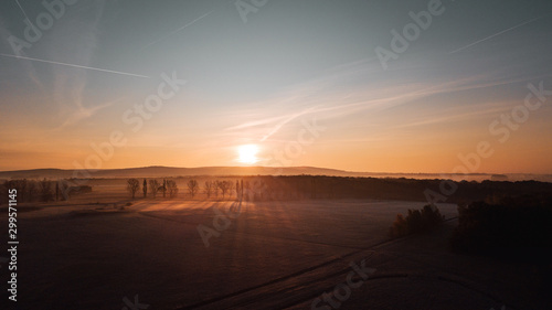 Aerial view from above of a countryside landscape with beautiful sunset light on a winter evening with forzen meadows and mist hazy horizon. Winter autumn trees with long shadows in germany