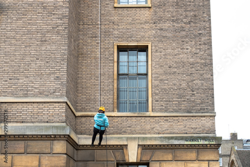 Young abseiling woman seen defending down a tall building in Cambridge city centre. She is wearing full protective gear, a team member can be seen atop the building.