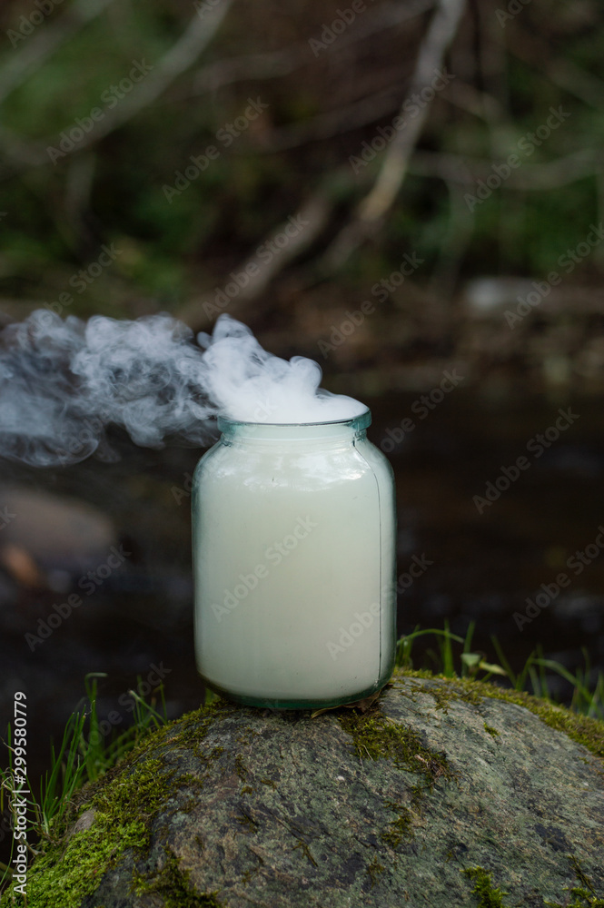 Glass jar full of smoke - magical view of smoke escaping from a jar - forest scene in the background a river Glass jar full of smoke - magical view of smoke escaping from a jar - forest scene in the background a river