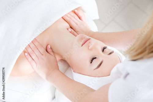 Young woman beautician doing neck and face massage to beautiful and charming young caucasian female client lying on couch under white blanket. Concept of cosmetic procedures and anti-aging products