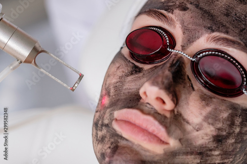 Close-up of a cosmetologist's hand making a hardware carbon peeling by a young pretty customer woman in safety glasses and a headdress lying on a couch in a beauty salon. Facial cleansing concept photo