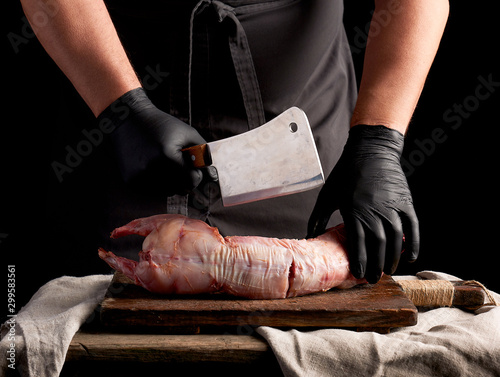 Photos chef in black latex gloves holds a big knife and cuts into pieces raw rabbit mea