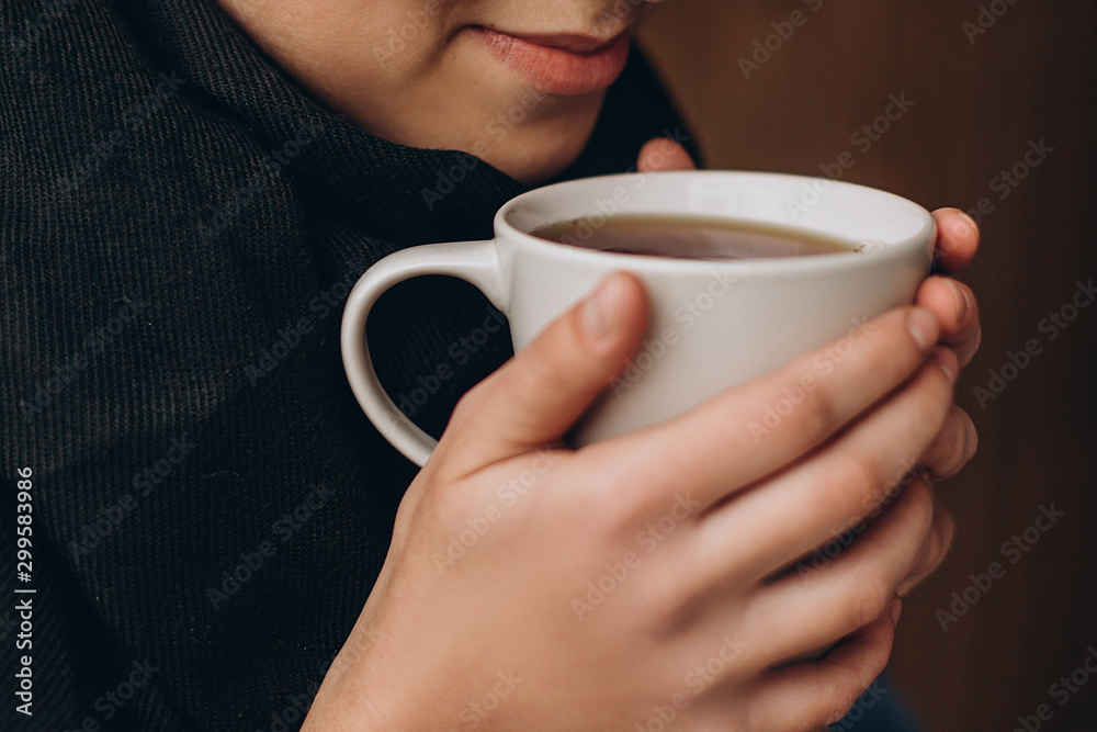 a mug of hot coffee or tea in the hands of a young pretty girl near her lips. Cropped photo with focus on cup