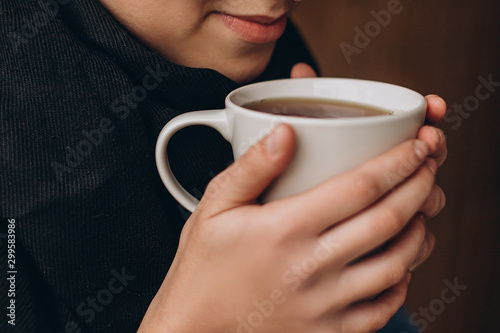 a mug of hot coffee or tea in the hands of a young pretty girl near her lips. Cropped photo with focus on cup