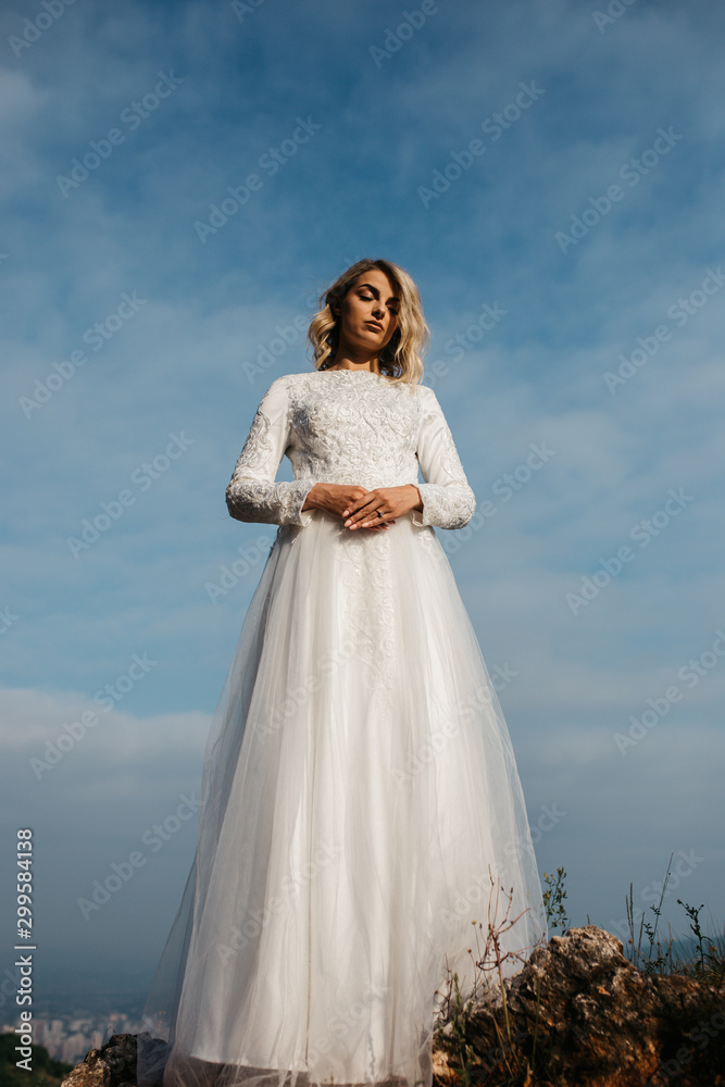 Beautiful elegant bride in lace wedding dress with long full skirt and long sleeves. Pretty girl in white. Nature, with city in the background.