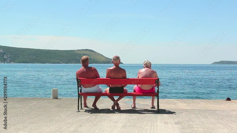 Old men sit on a bench by the sea in summer. Three old buddies sunbathe on the bench in the summer Old men sit on a bench by the sea in summer. Three old buddies sunbathe on the bench in the summer