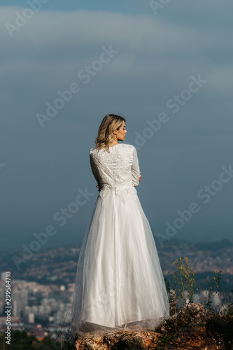 Beautiful elegant bride in lace wedding dress with long full skirt and long sleeves. Pretty girl in white. Nature, with city in the background.