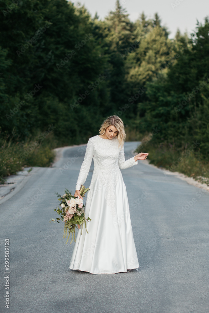 Beautiful elegant bride in lace wedding dress with long full skirt and long sleeves. She is holding a big bouquet of flowers. Outdoors, on the road. Beautiful elegant bride in lace wedding dress with long full skirt and long sleeves. She is holding a big bouquet of flowers. Outdoors, on the road.